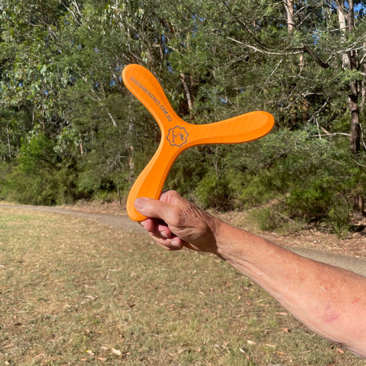 OZzie Emu Orange boomerang held by a person in an outdoor setting with trees in the background